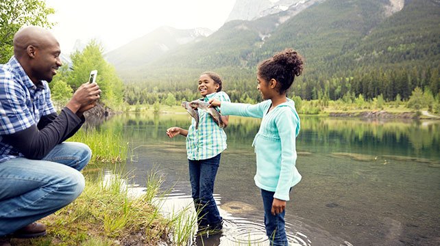 dad taking picture of daughters holding fish by a lake and smiling