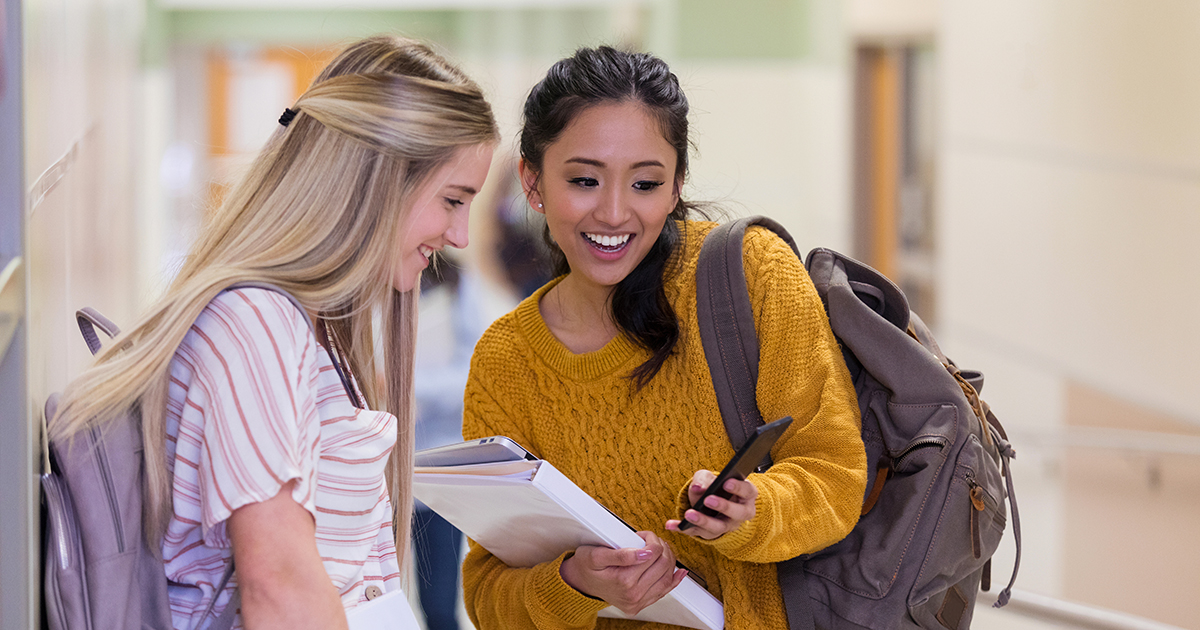 two high school girls walk through hallway smiling looking at cell phone