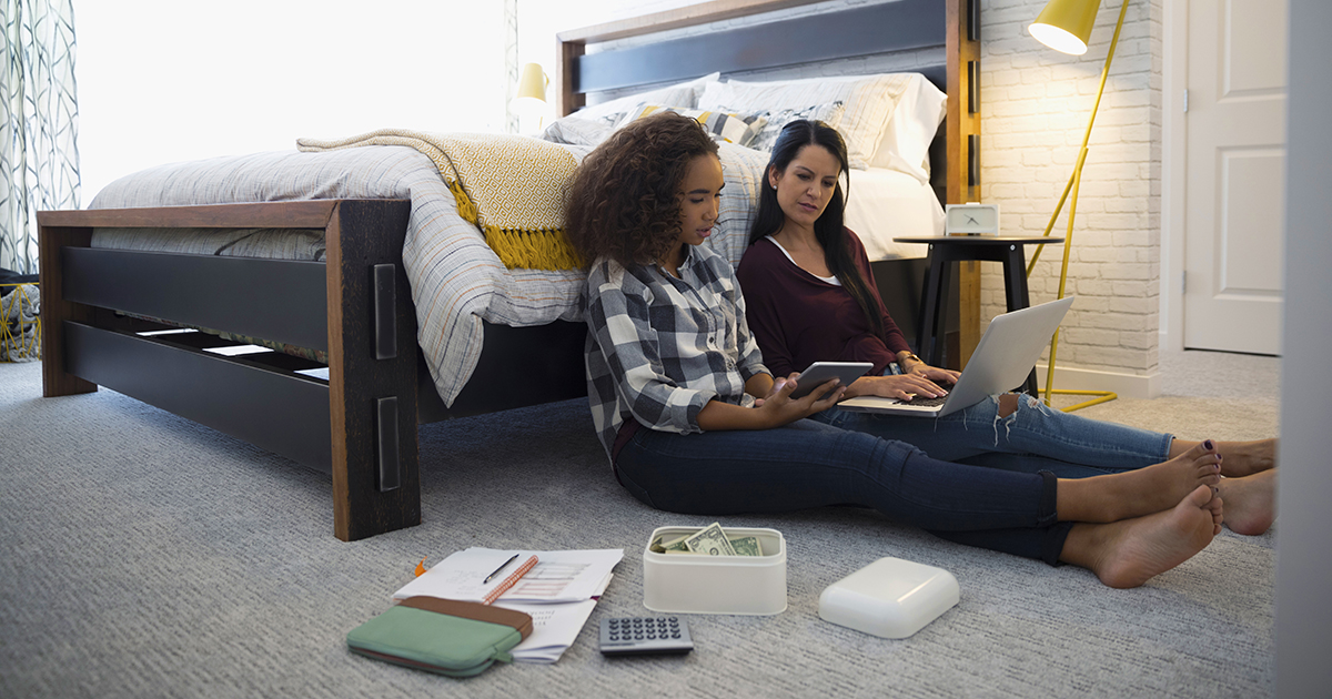 mom and daughter sit on bedroom floor and loop at laptop with calculator and money beside them
