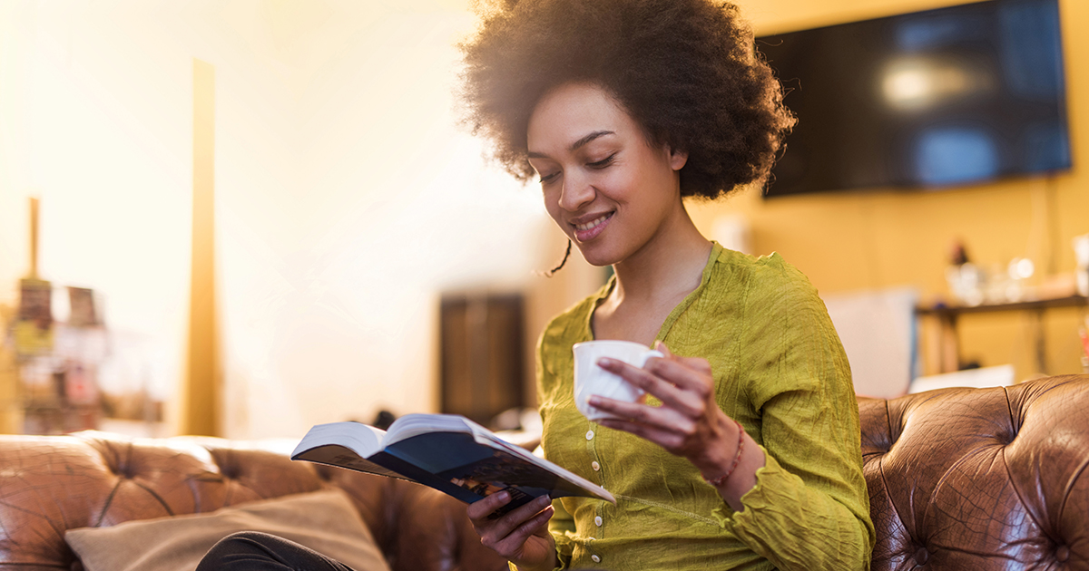 young woman drinks coffee and reads book on couch in living room